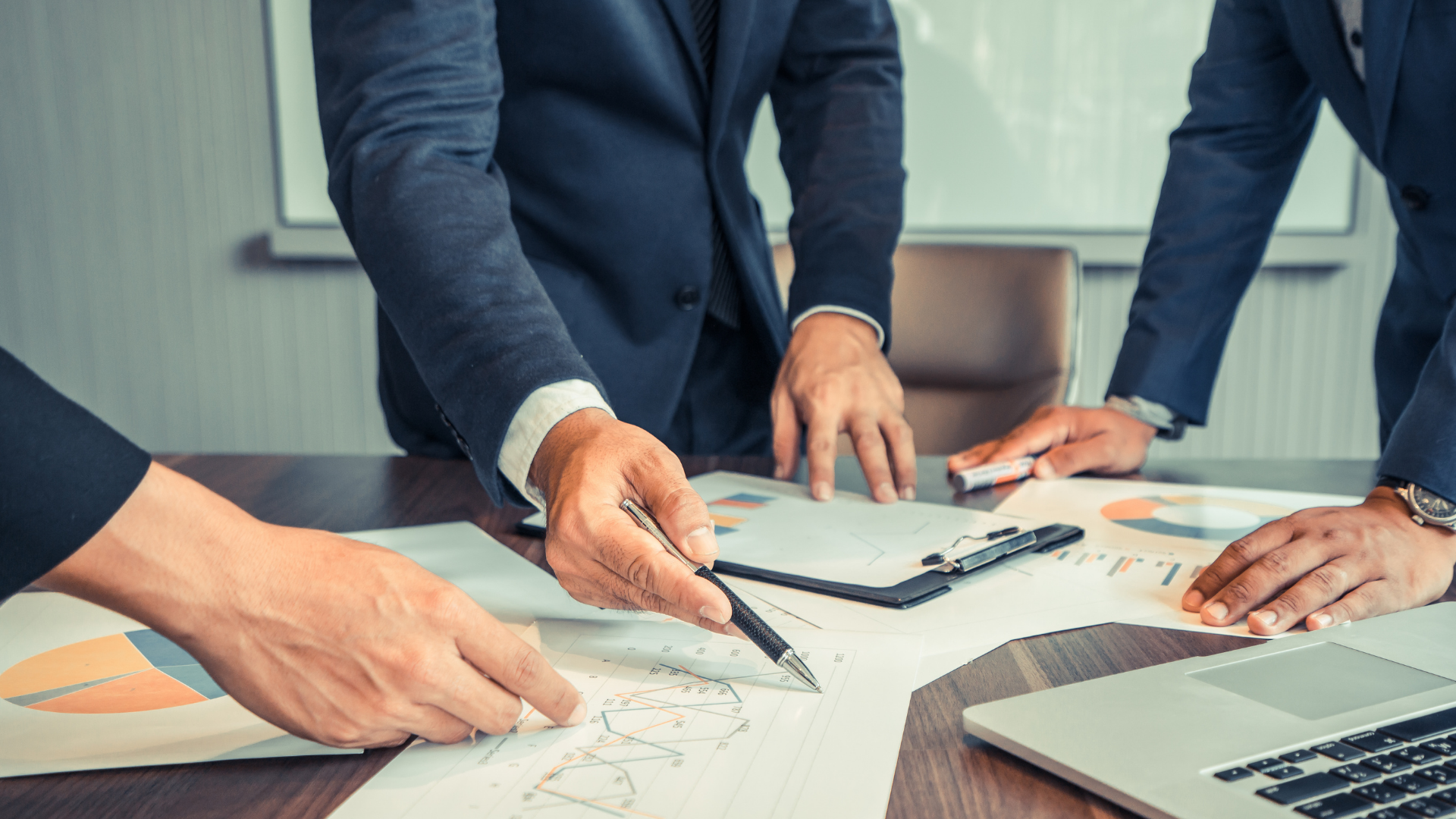 Hands of businesspeople reviewing documents and pointing at charts around a laptop in a meeting room