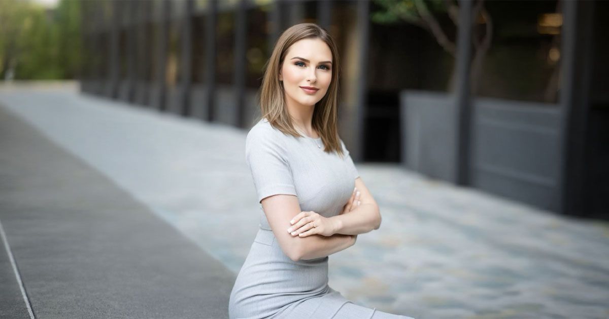 A person in a gray dress sits with arms crossed outside a modern building with a paved walkway.