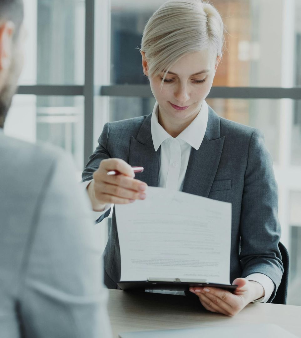 A person in a business suit presents a document on a clipboard to someone sitting across from them in an office setting.