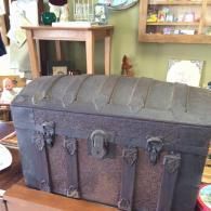 A dark, vintage, rounded-top trunk with metal latch hardware, sitting on a wooden surface in a shop.