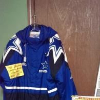 A blue, white, and black Dallas Cowboys jacket hangs on a hanger against a wooden door, featuring a yellow price tag.
