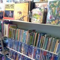 A wooden shelf displays vinyl records, books, and DVDs arranged in rows inside a store.