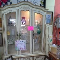 A cream-colored, three-door wooden china cabinet with glass panels displaying various decorative ceramic items inside.