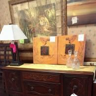 A wooden sideboard topped with a lamp and two decorative art pieces, positioned in front of a framed landscape painting.