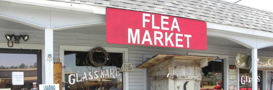 A red sign reading "FLEA MARKET" hangs above the entrance of a shop with glass windows labeled "GLASSWARE" and "GIFTS".