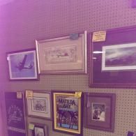 A collection of framed artwork and photographs displayed on a beige pegboard wall.