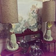 Two table lamps featuring pale green hand-shaped bases with gold-painted cuffs sit on a table, flanked by metallic items.