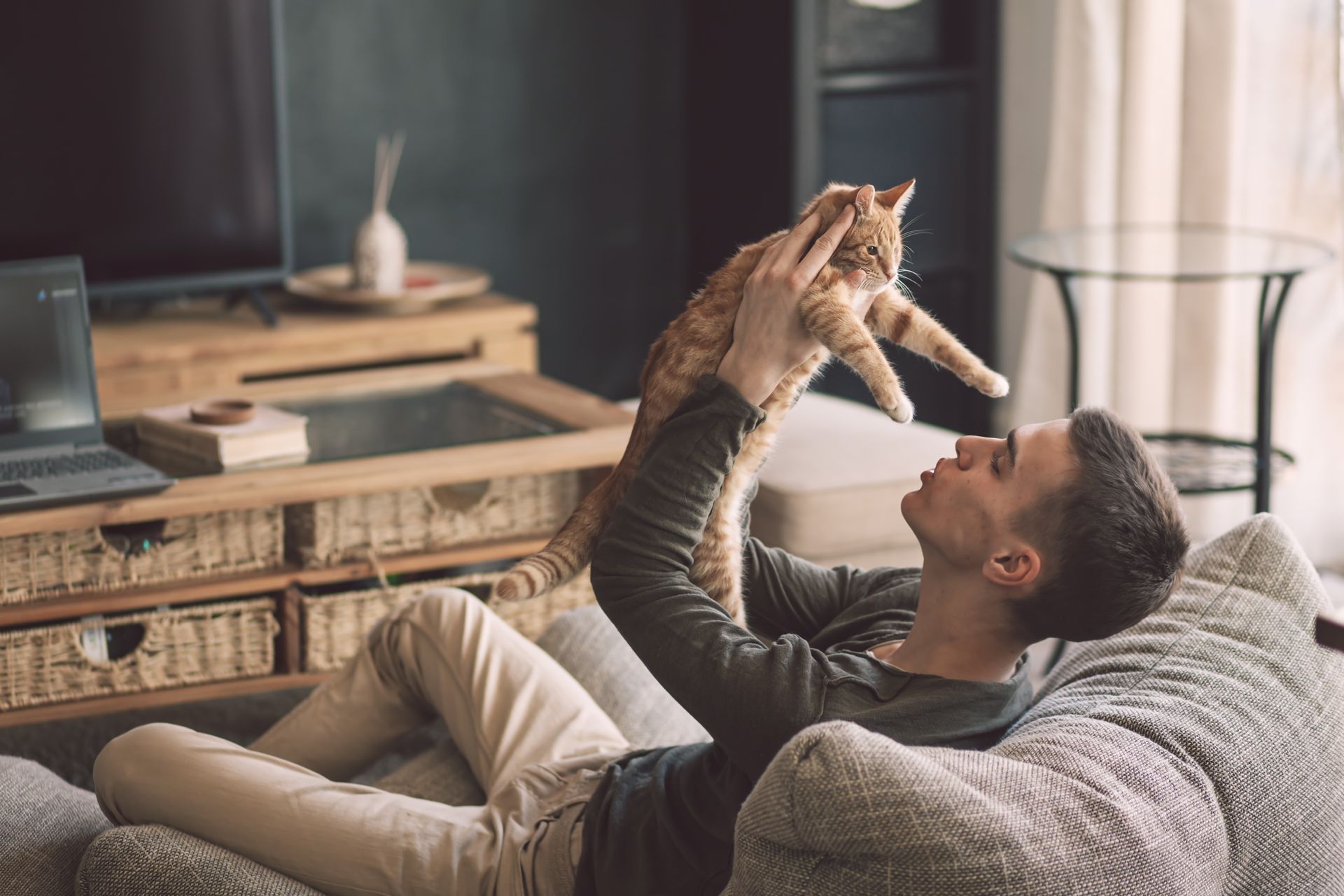 Man lying on couch holding orange tabby cat up, smiling. Living room setting.