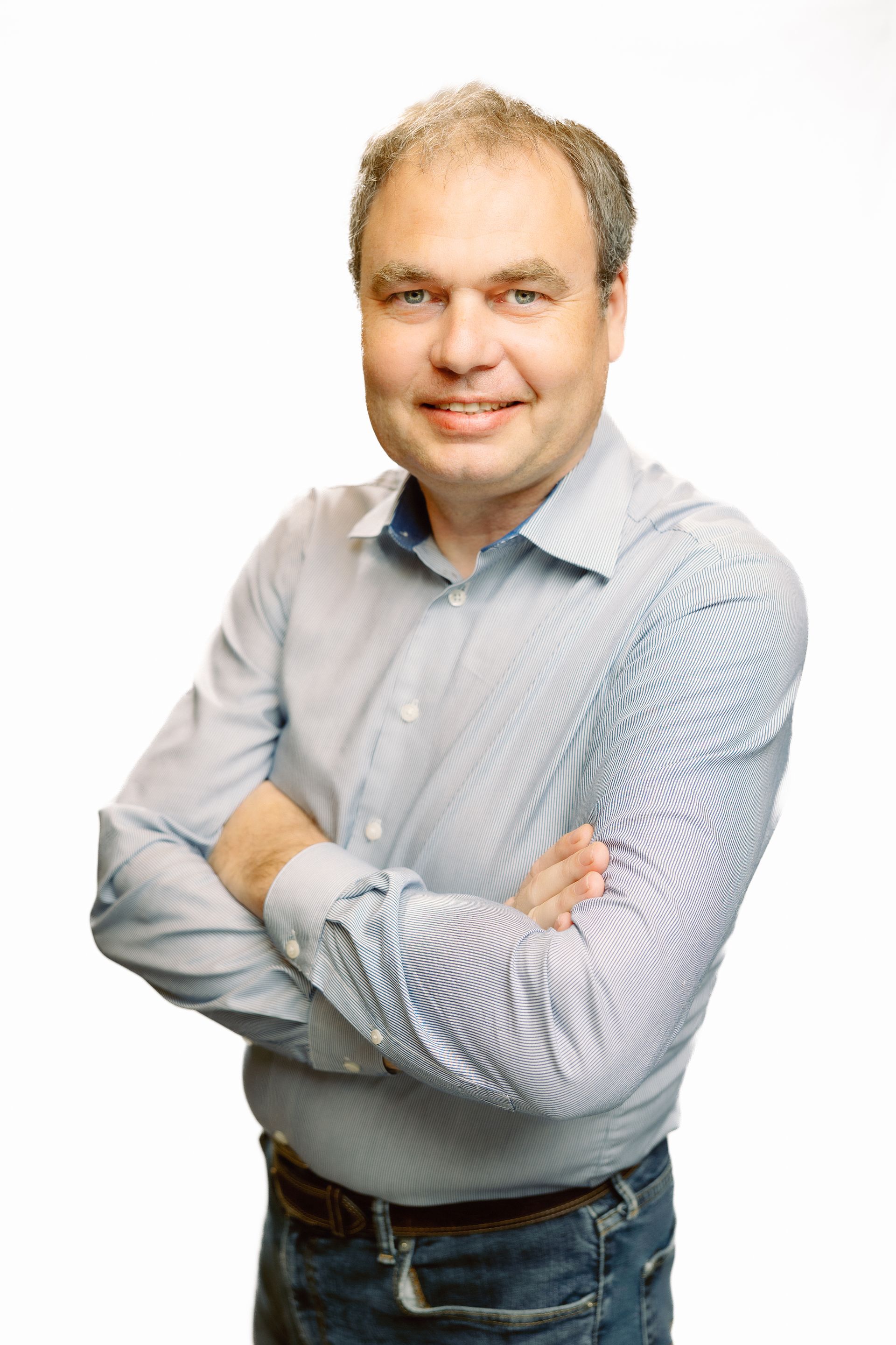 Man with arms crossed, smiling, wearing a blue button-down shirt and jeans, against a white background.