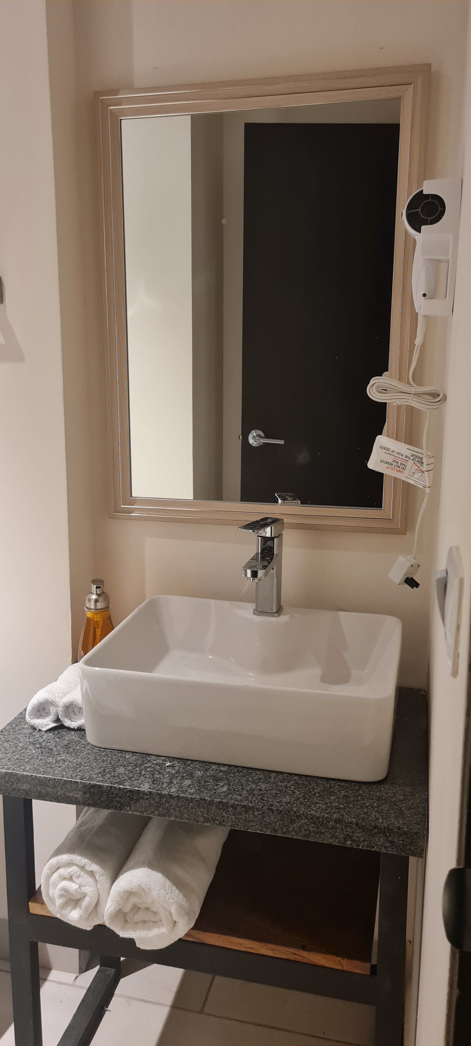 Bathroom sink with white basin, rectangular mirror, and rolled towels on a shelf.