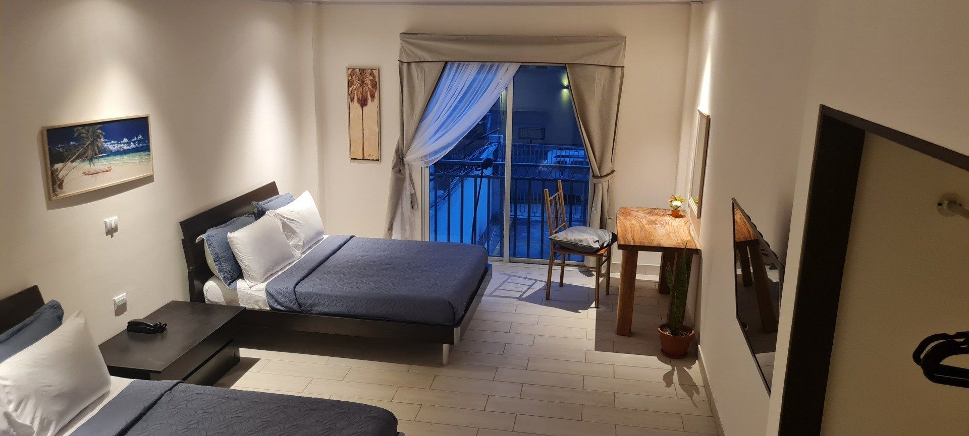 A furnished room with two beds, a small table, and a balcony. The room has soft lighting and neutral tones.