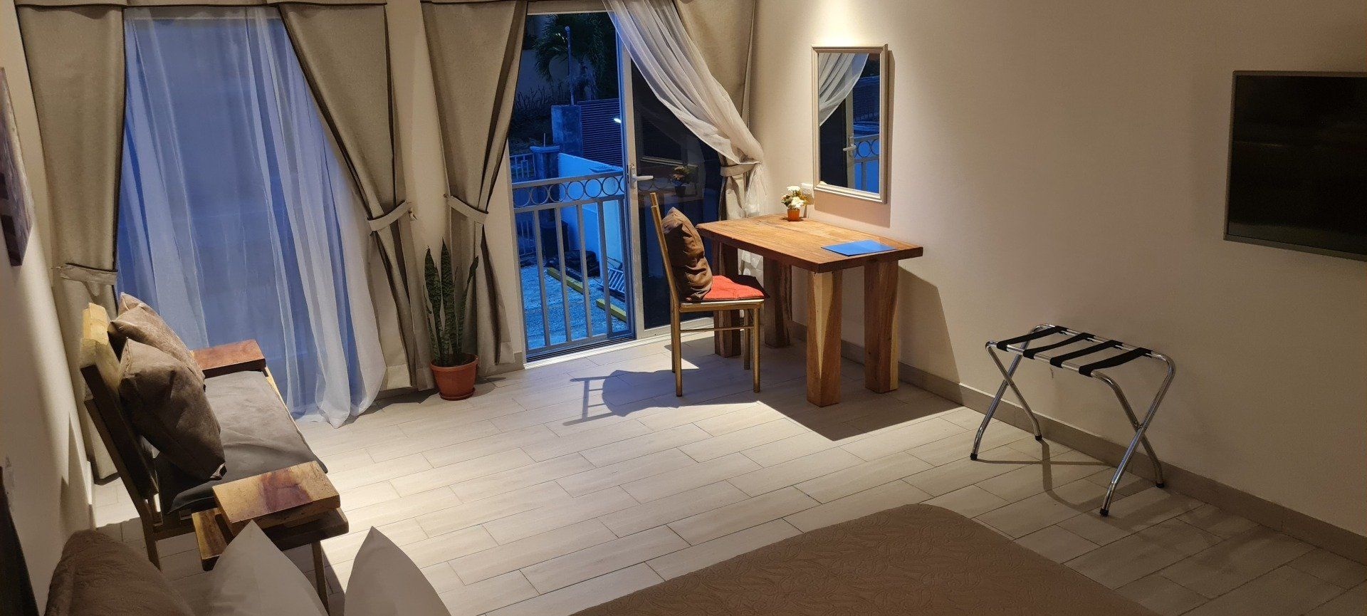A brightly lit room with a table, chair, and luggage rack in front of a window. A TV is on the wall.