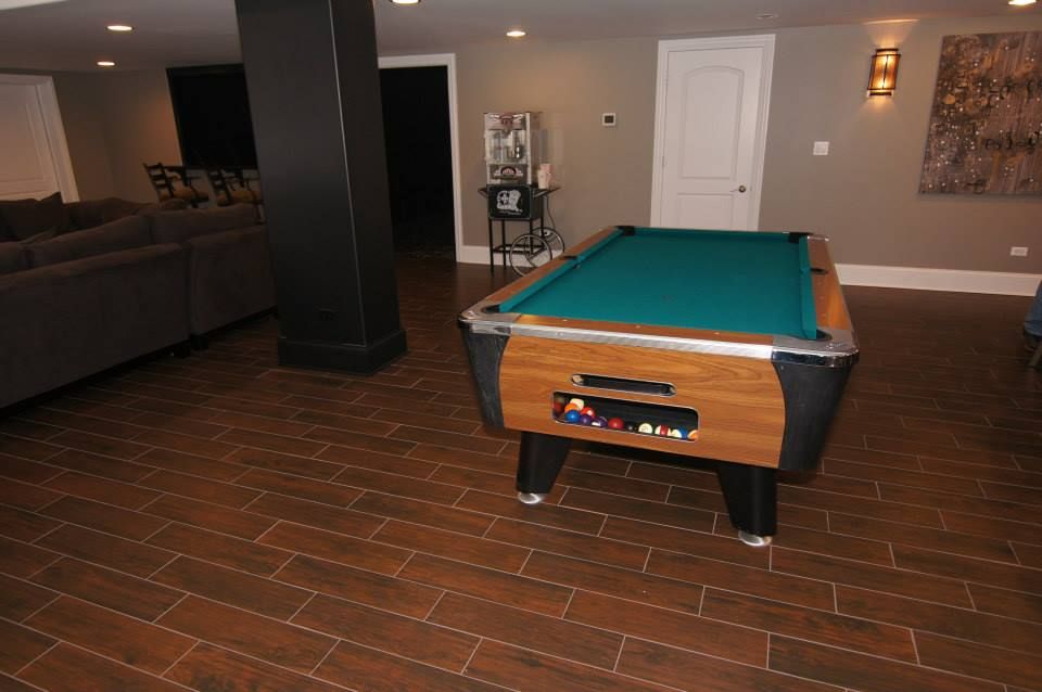 A basement recreation room featuring a pool table with a green felt surface, wood-tone sides, and dark tile flooring.