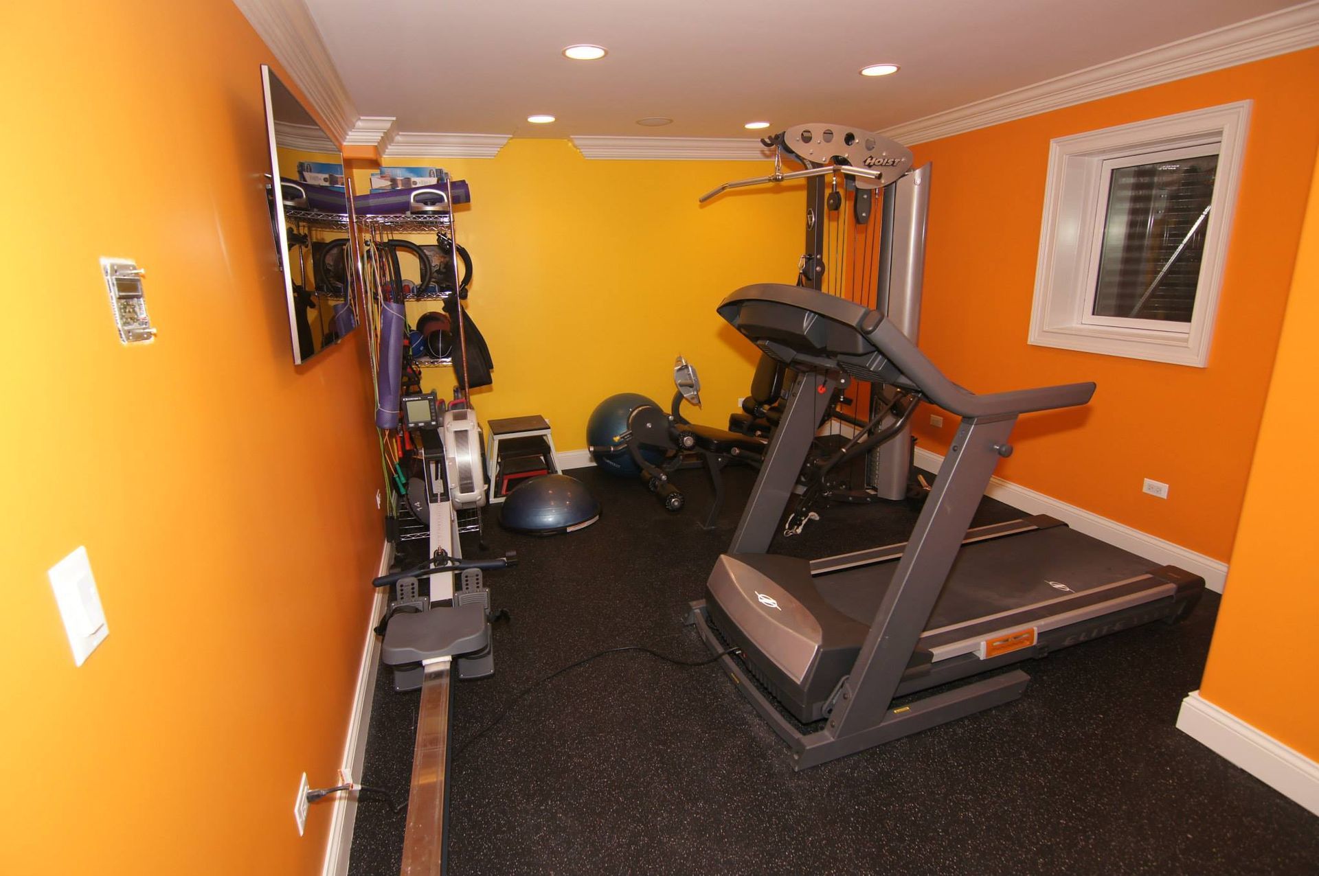 Home gym with orange walls, rubber flooring, a treadmill, a rowing machine, and various fitness equipment.