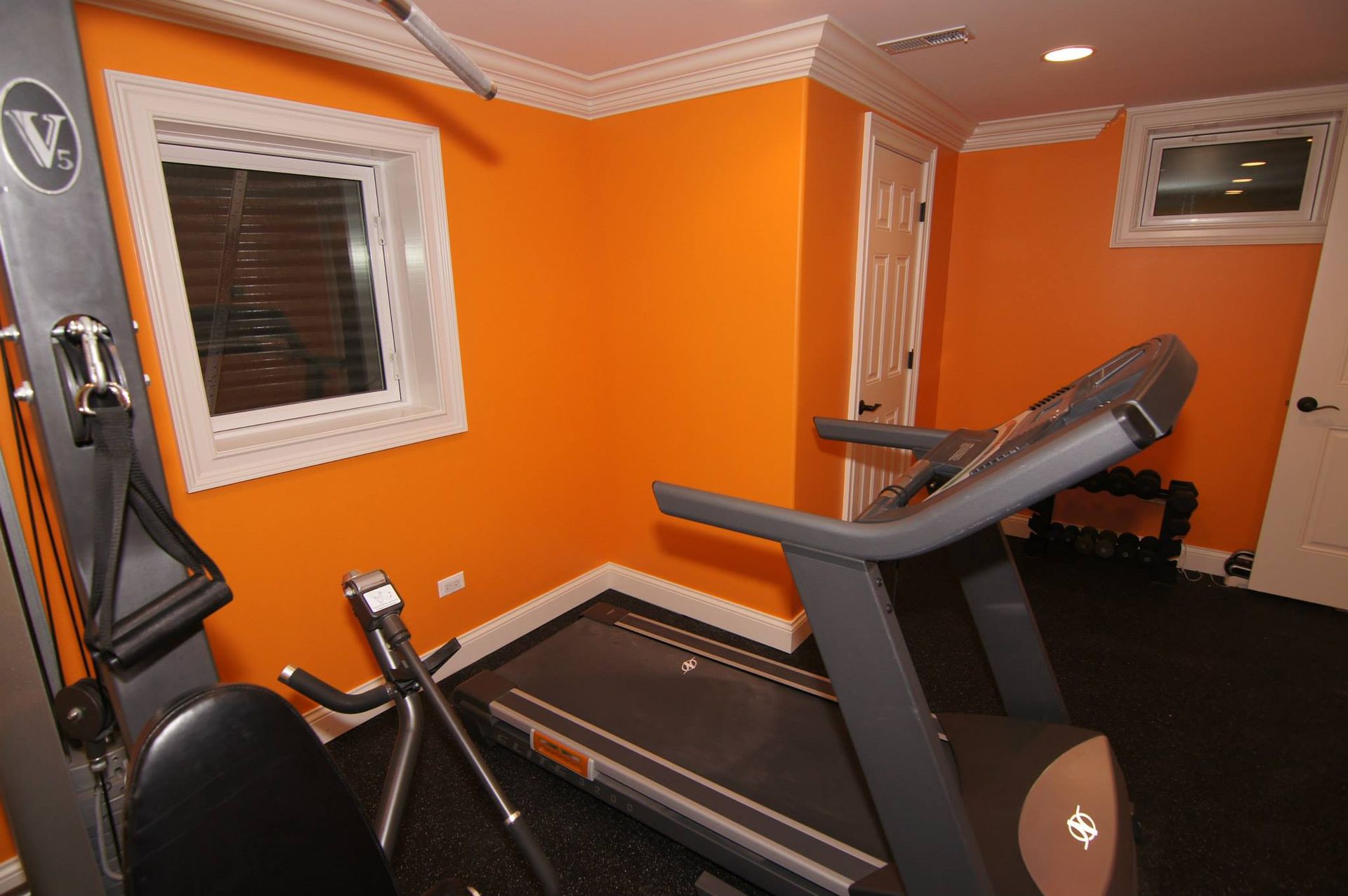A home gym with orange walls, a treadmill, a cable machine, and a small dumbbell rack on a black floor.