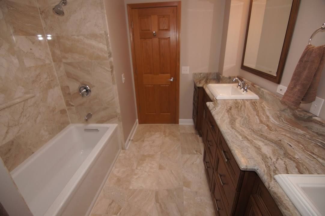 A bathroom with light stone tile flooring, a shower-tub combo with tiled walls, and a wood vanity with a marble countertop.