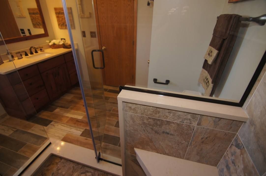 A bathroom featuring a wood-look tile floor, a wooden vanity with a white countertop, and a walk-in shower with stone tiles.