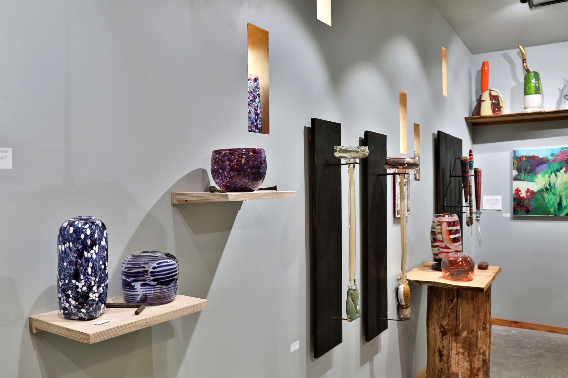 Gallery wall with artwork: glass sculptures, paintings, wooden shelves against a gray wall.