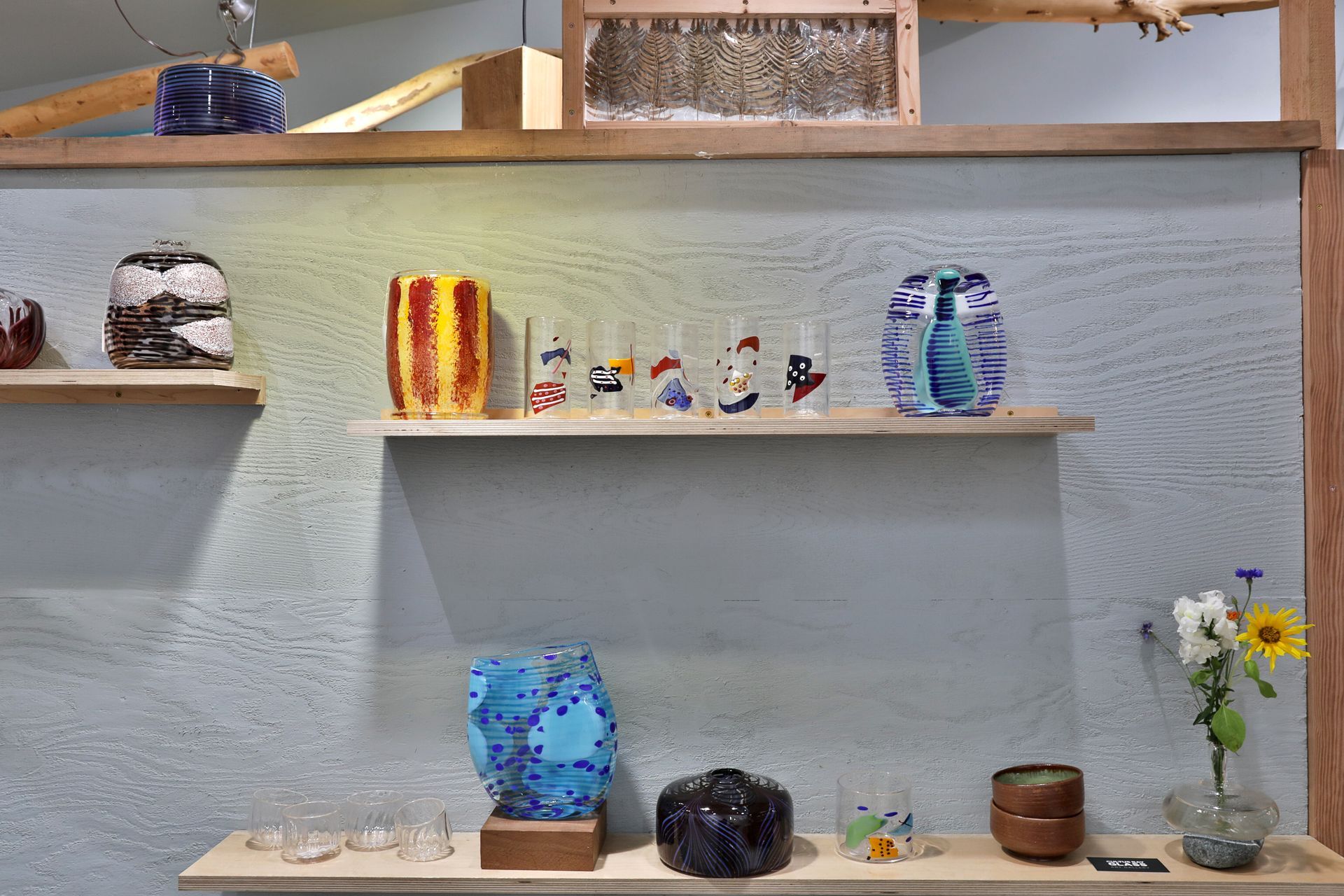 Shelves display colorful glass art in a shop against a textured grey wall.