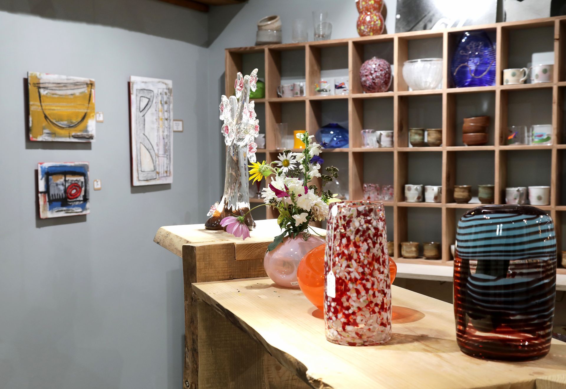 Art gallery interior: artworks on wall, handcrafted glass and ceramics on display shelf and counter.