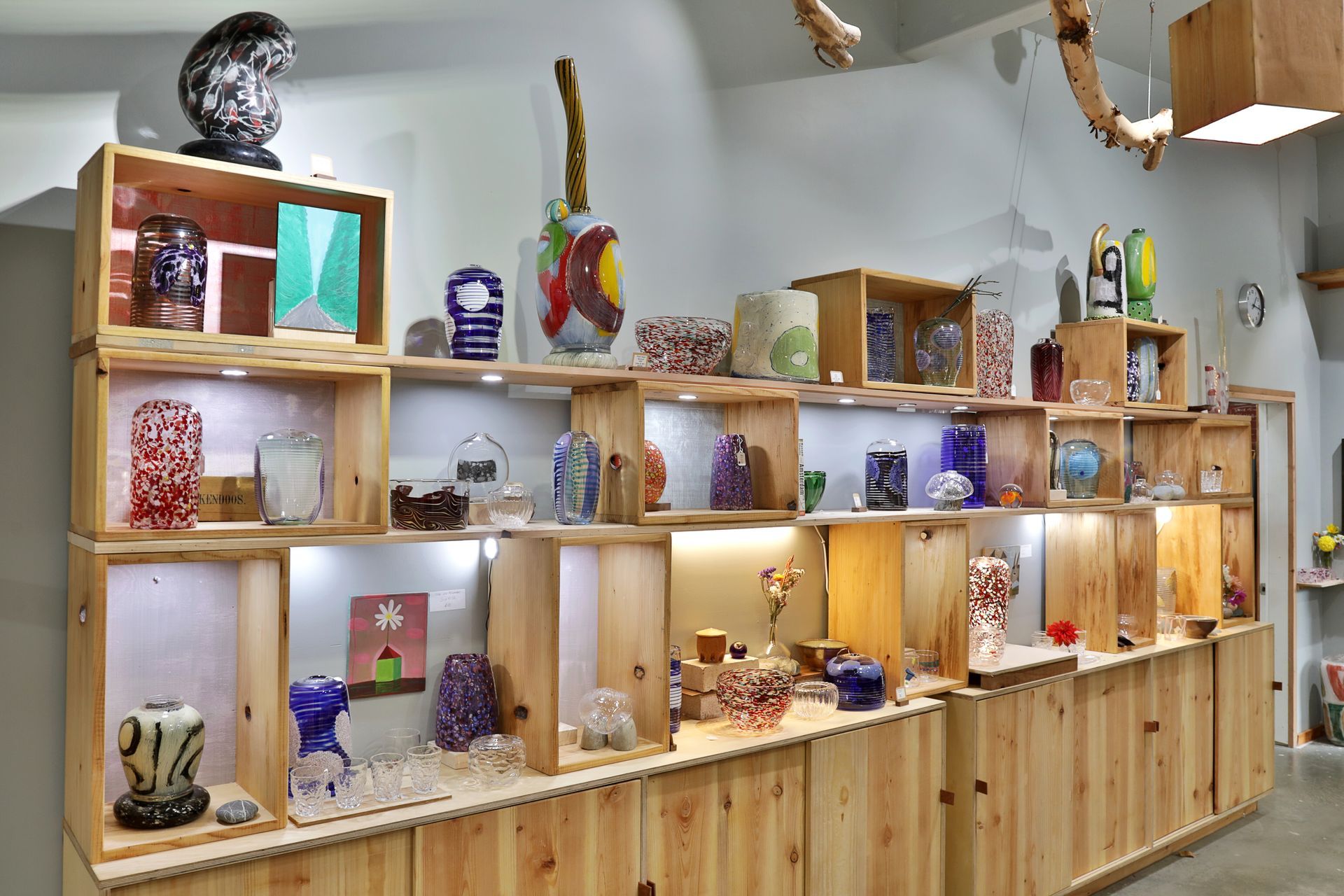 Wooden shelves displaying various colorful glass art pieces in a well-lit gallery space.