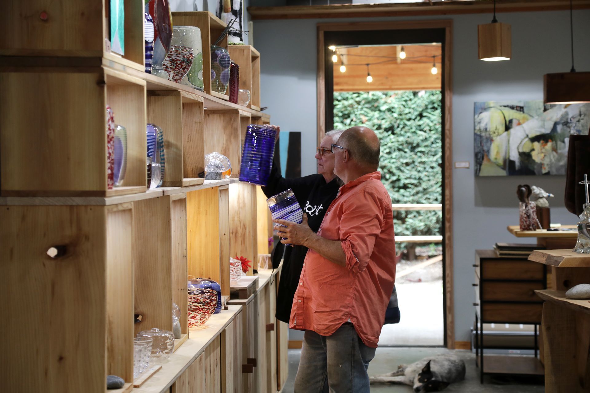 Two people browsing glass art in a gallery; one holds a vase. Wooden shelves line the wall.