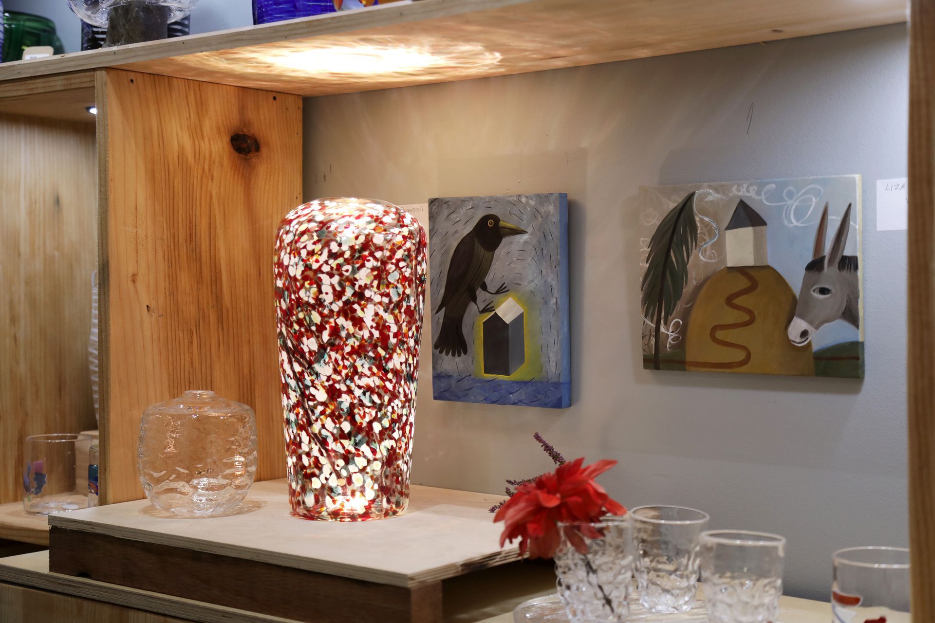 Glassware display: A speckled red and white vase illuminated, with two paintings of a crow and a house, and clear glasses.