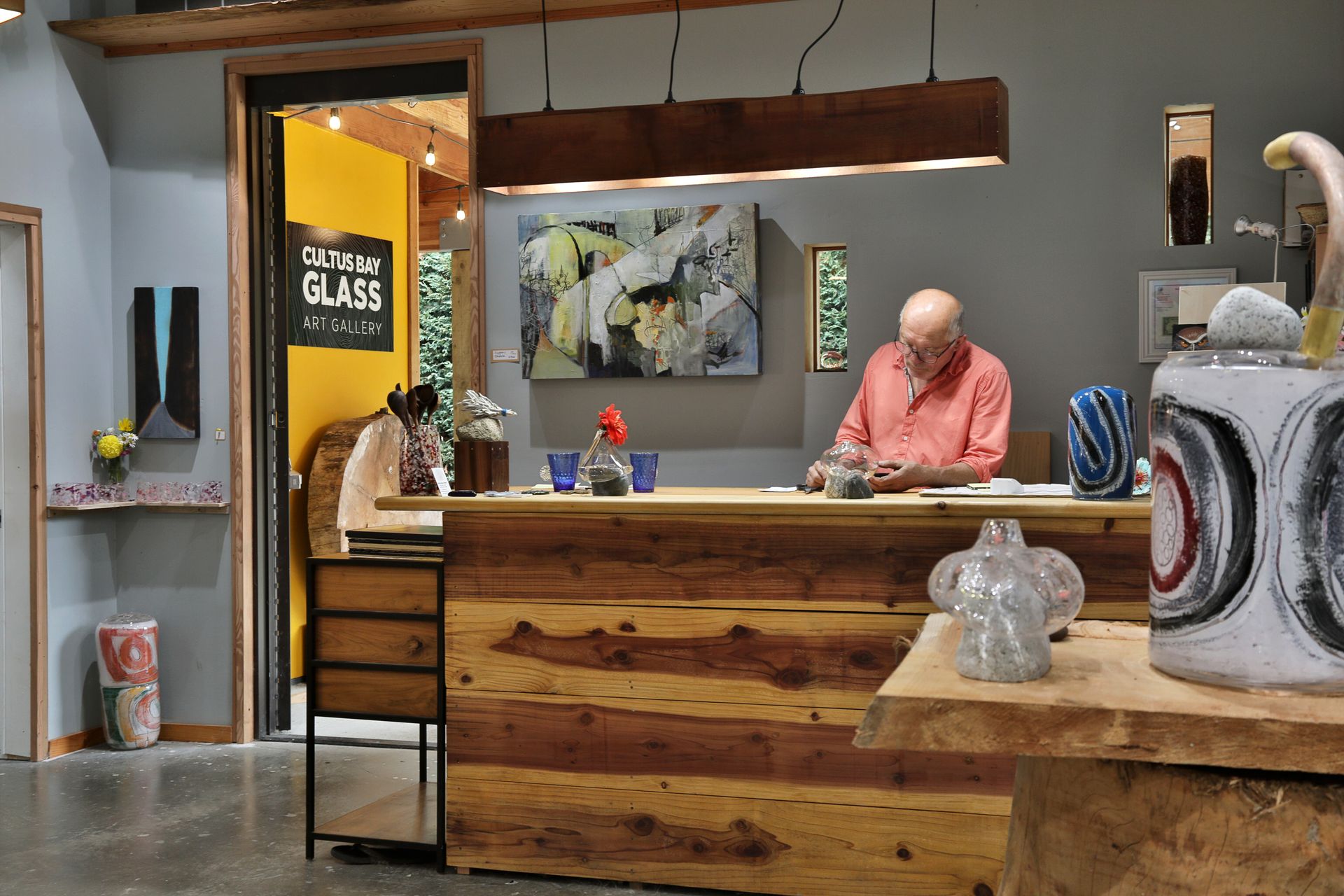 Man at a wooden counter in a glass art studio; studio setting with art pieces and a yellow door.