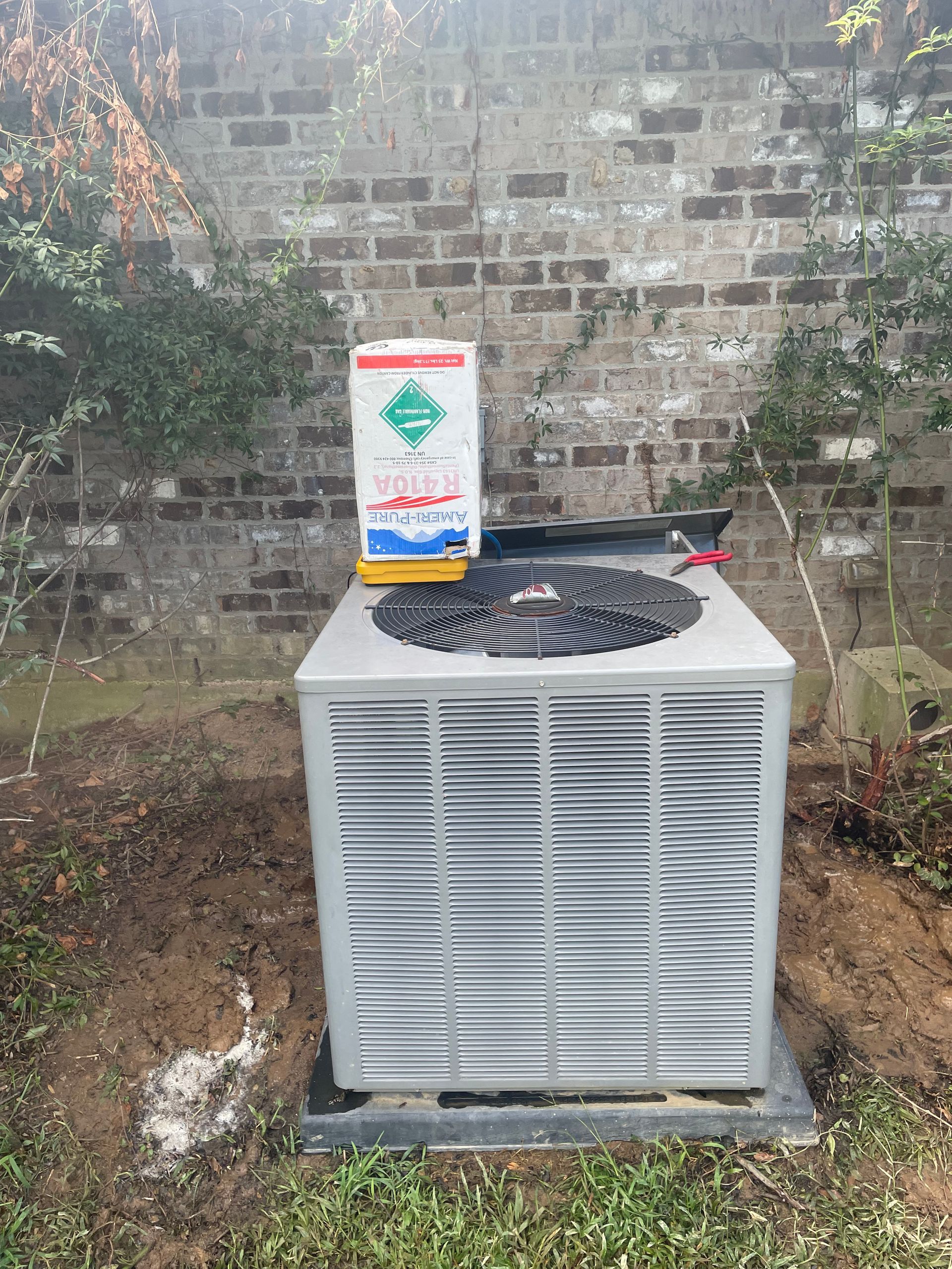 An air conditioner is sitting in the grass next to a brick wall.