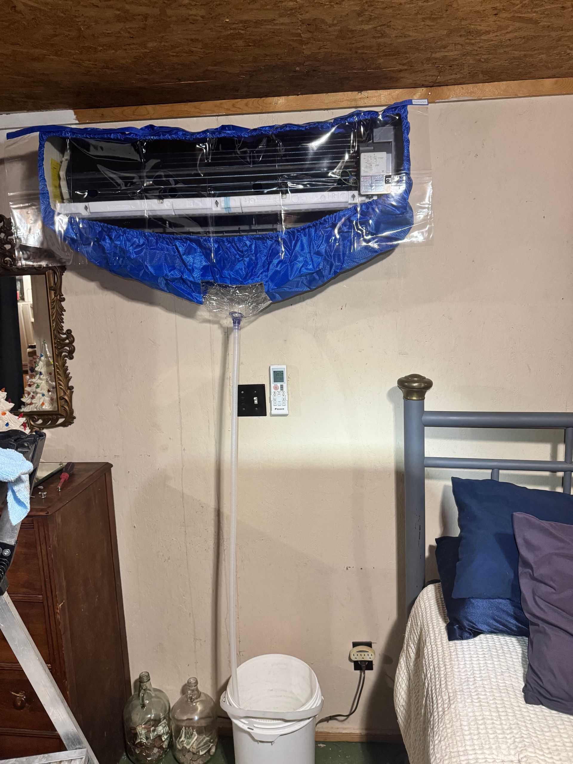 A room with a bed , dresser , trash can and air conditioner.