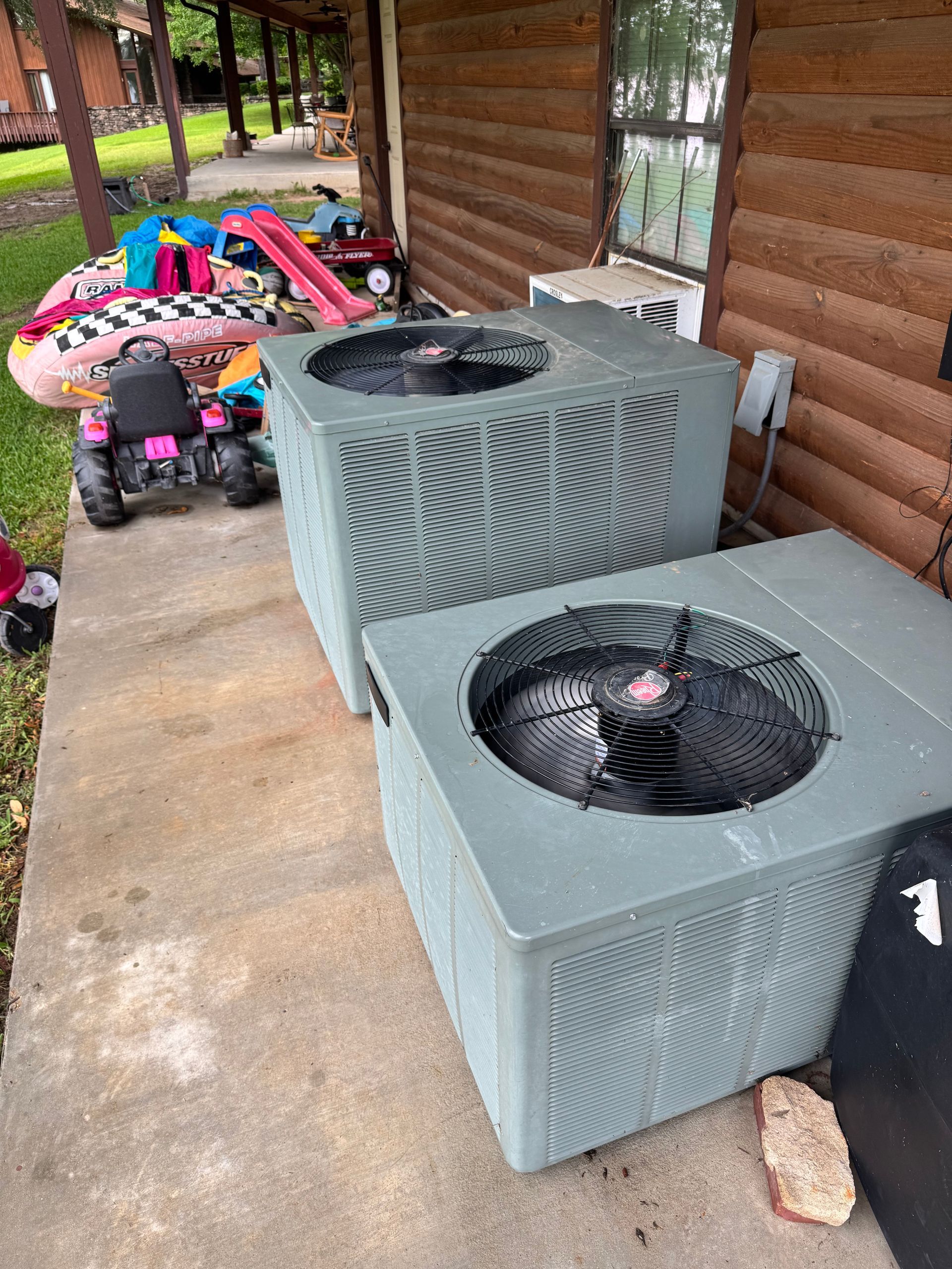 Two air conditioners are sitting on a porch next to a log cabin.