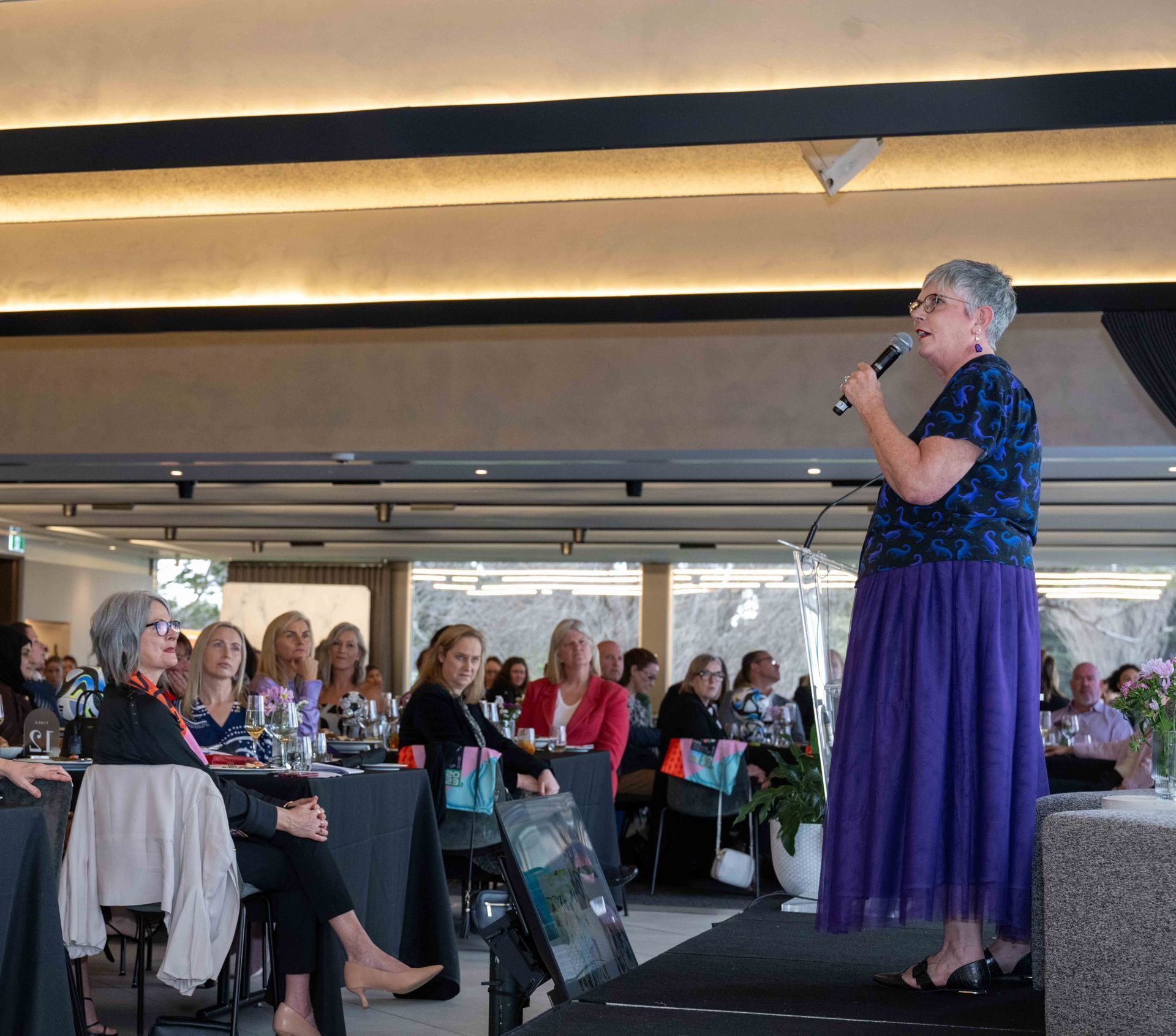 Woman speaking at event; purple skirt, microphone, audience seated at tables.