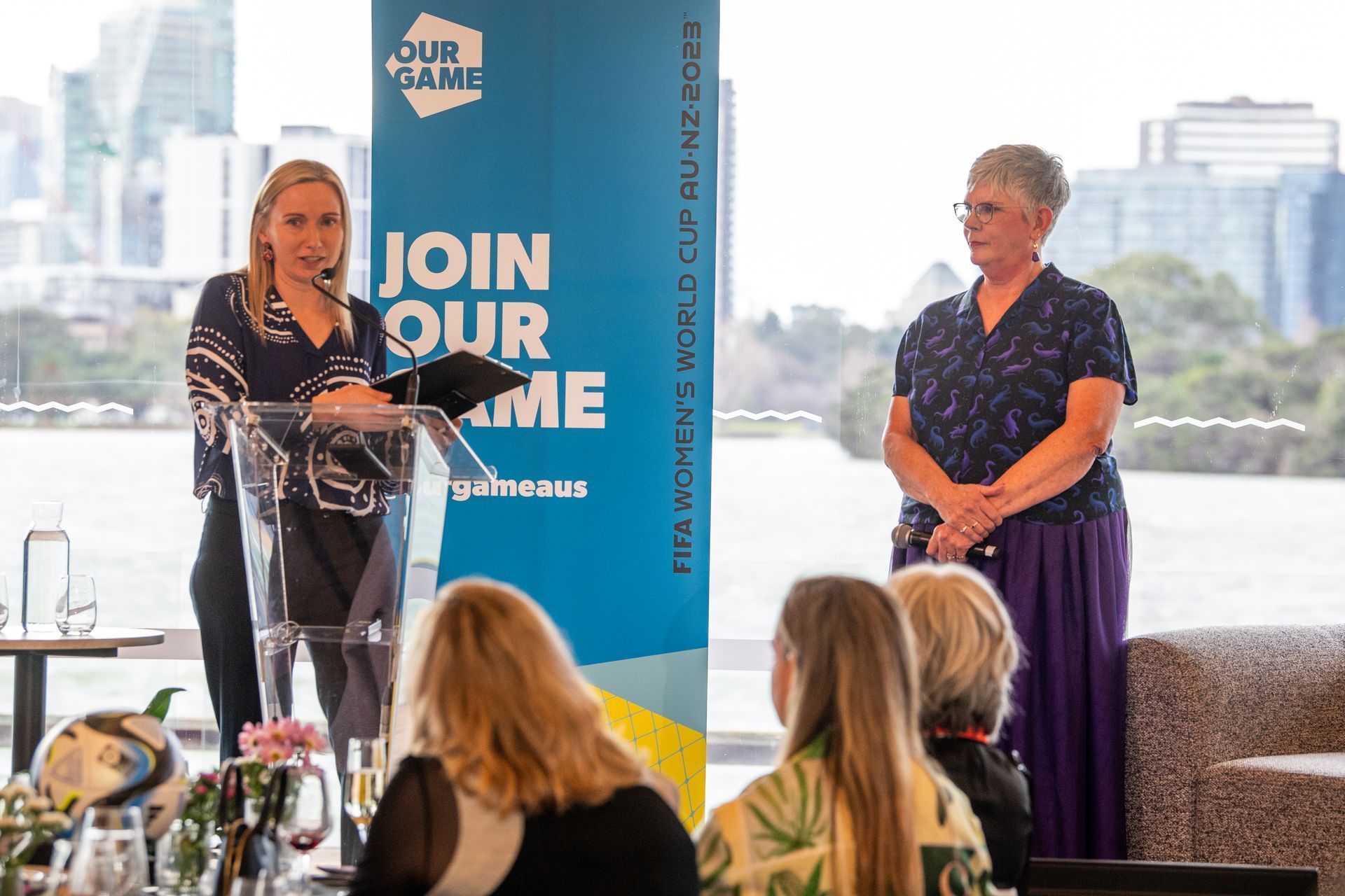 Two women at a podium speaking to an audience. A banner reads 