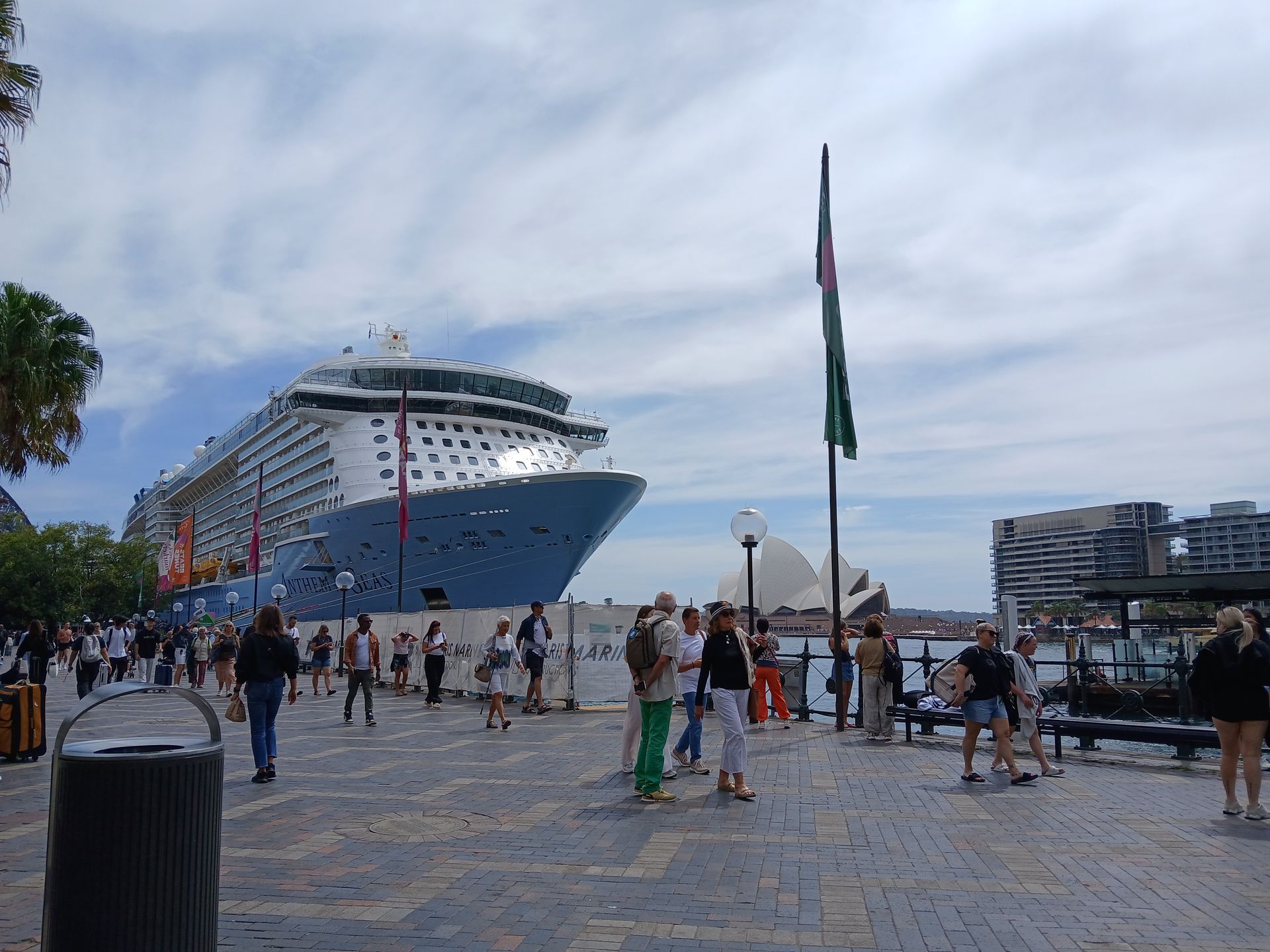 Large cruise ship docked at a harbor, people walking on the pier under a cloudy sky.