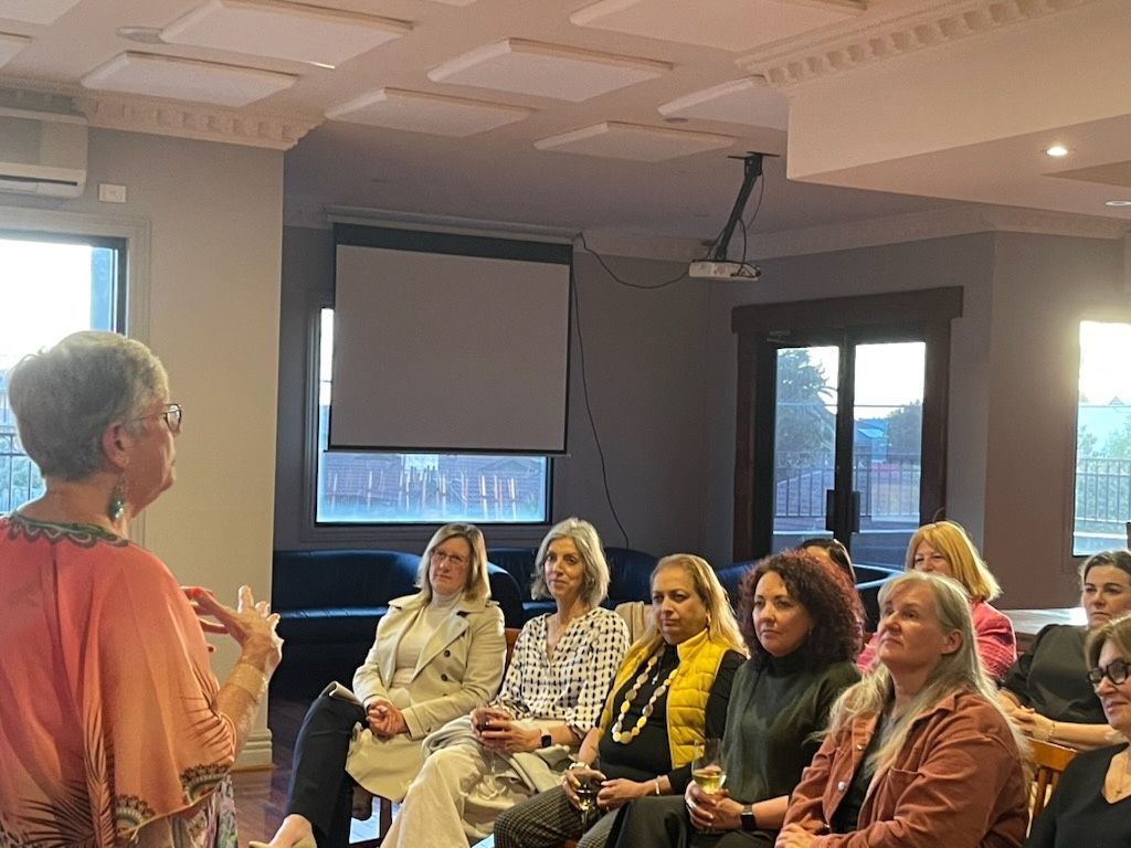 Woman speaking to a seated group. Audience listens attentively in a room with a projector screen.