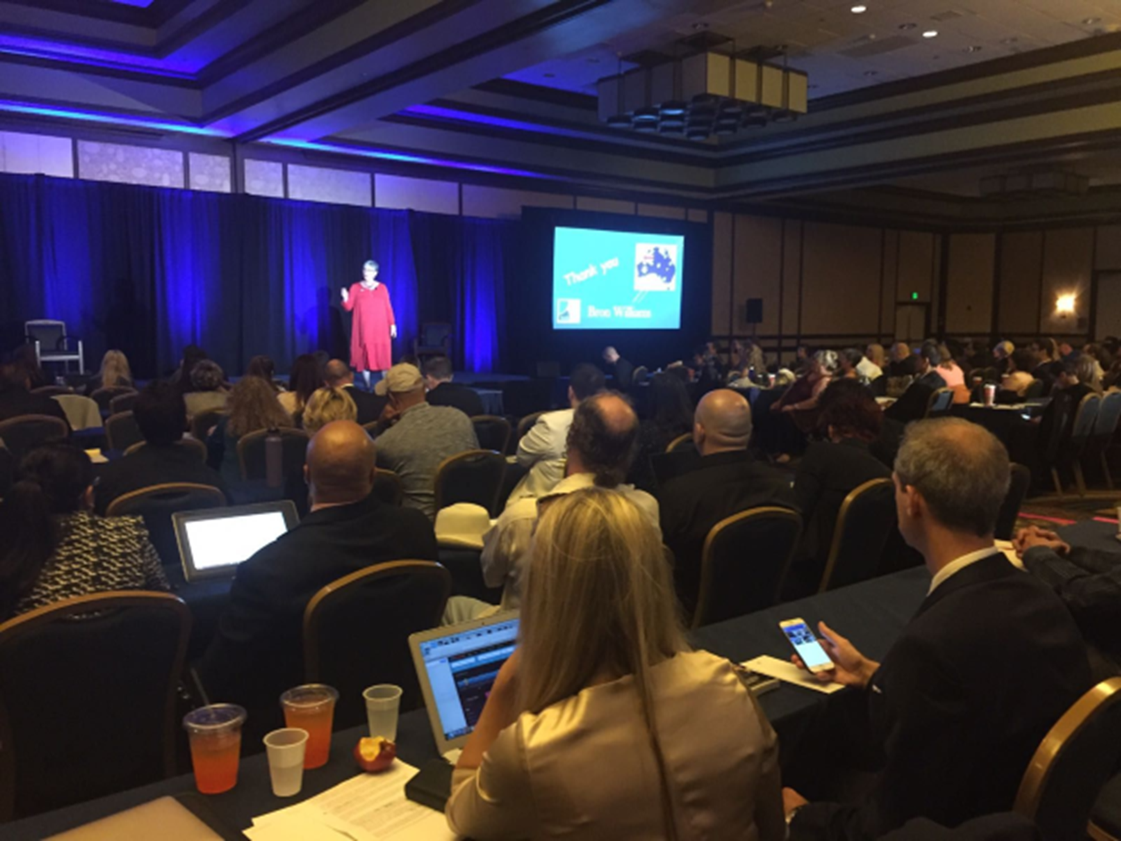 Audience watches speaker on stage at a conference. The speaker wears a red dress. Blue lights illuminate the stage.