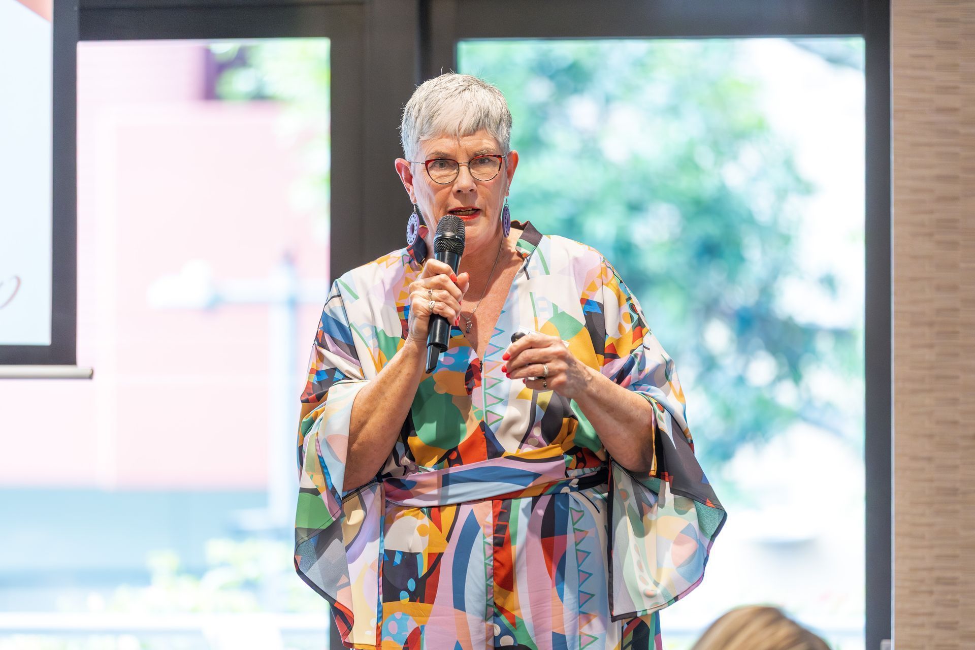 Woman speaking into a microphone, wearing a colorful patterned robe, in a room with a window.