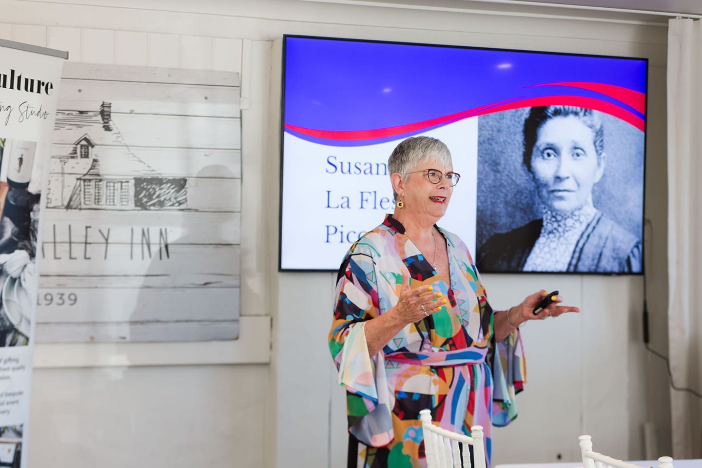 Woman presenting, with a photo of Susan La Flesche Picotte on a screen; an Inn sign is to the left.