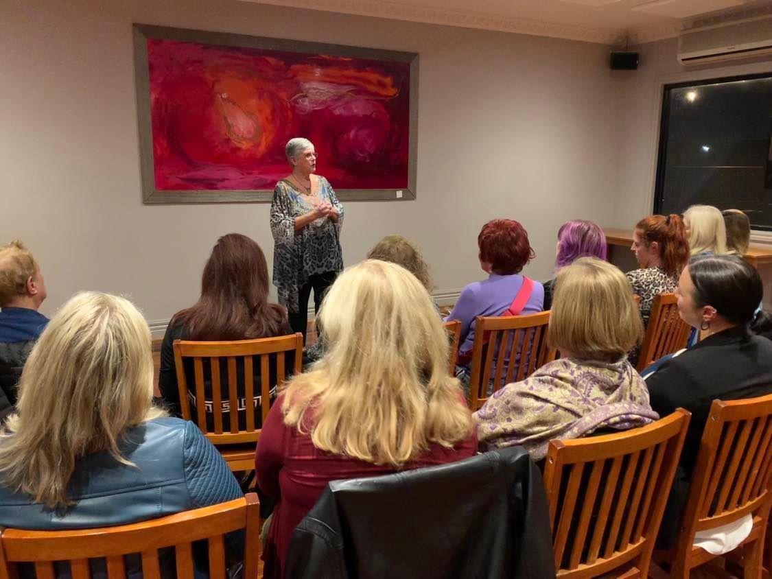 Woman speaking to an audience seated in wooden chairs; abstract red painting on the wall.