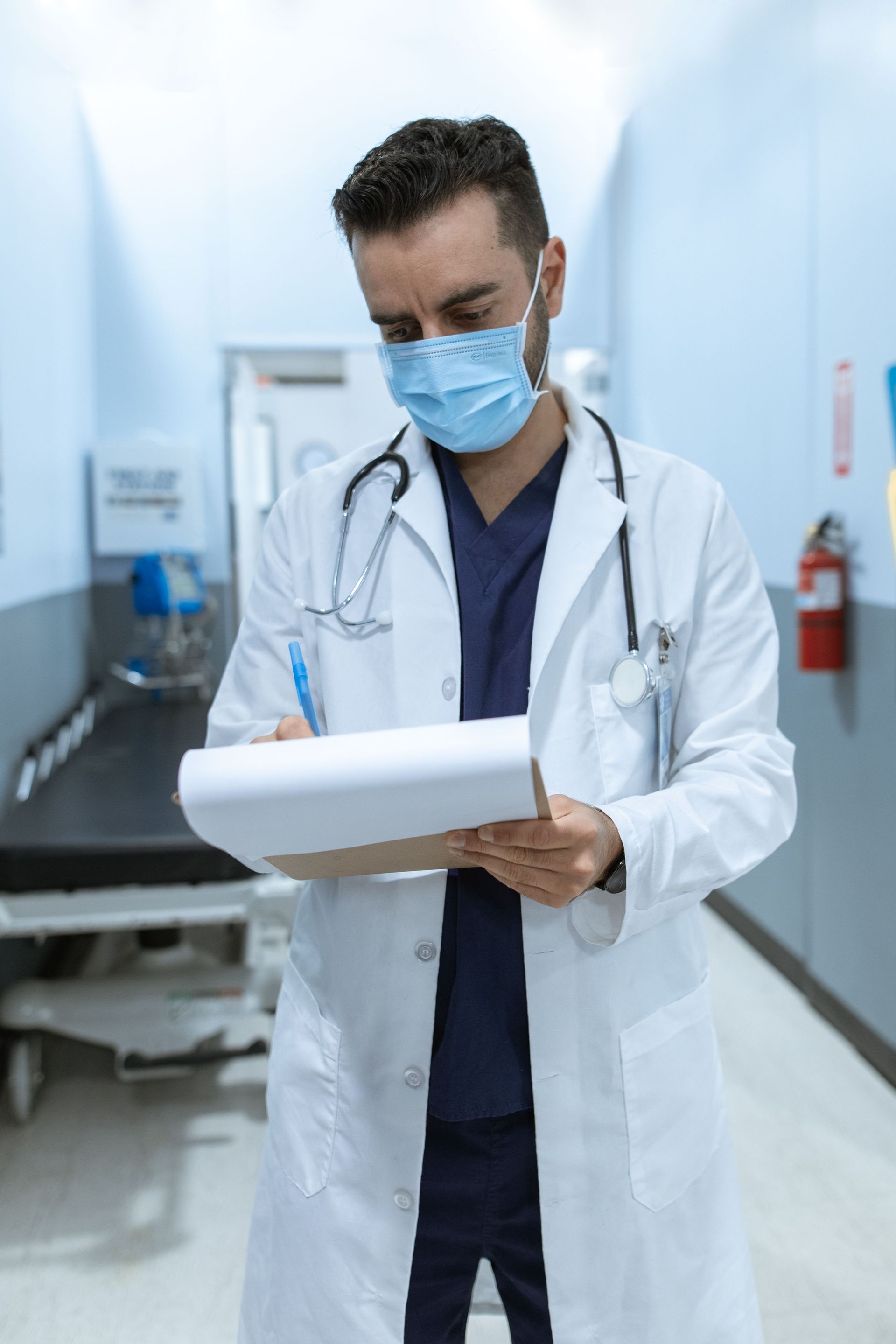 Doctor wearing a mask and lab coat reviewing a clipboard in a hospital corridor