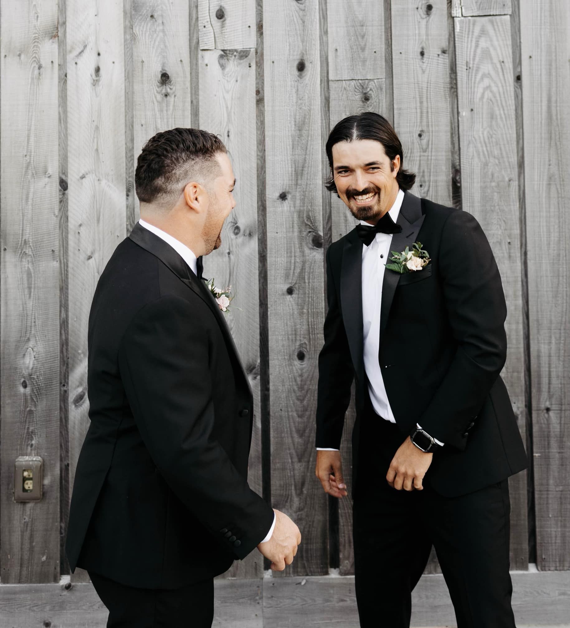 Two men in tuxedos are laughing in front of a wooden wall.