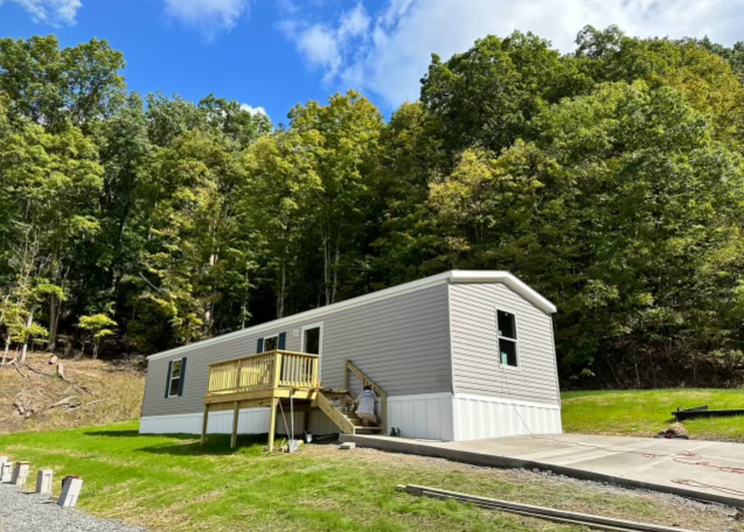 A mobile home with a deck in the middle of a field surrounded by trees.