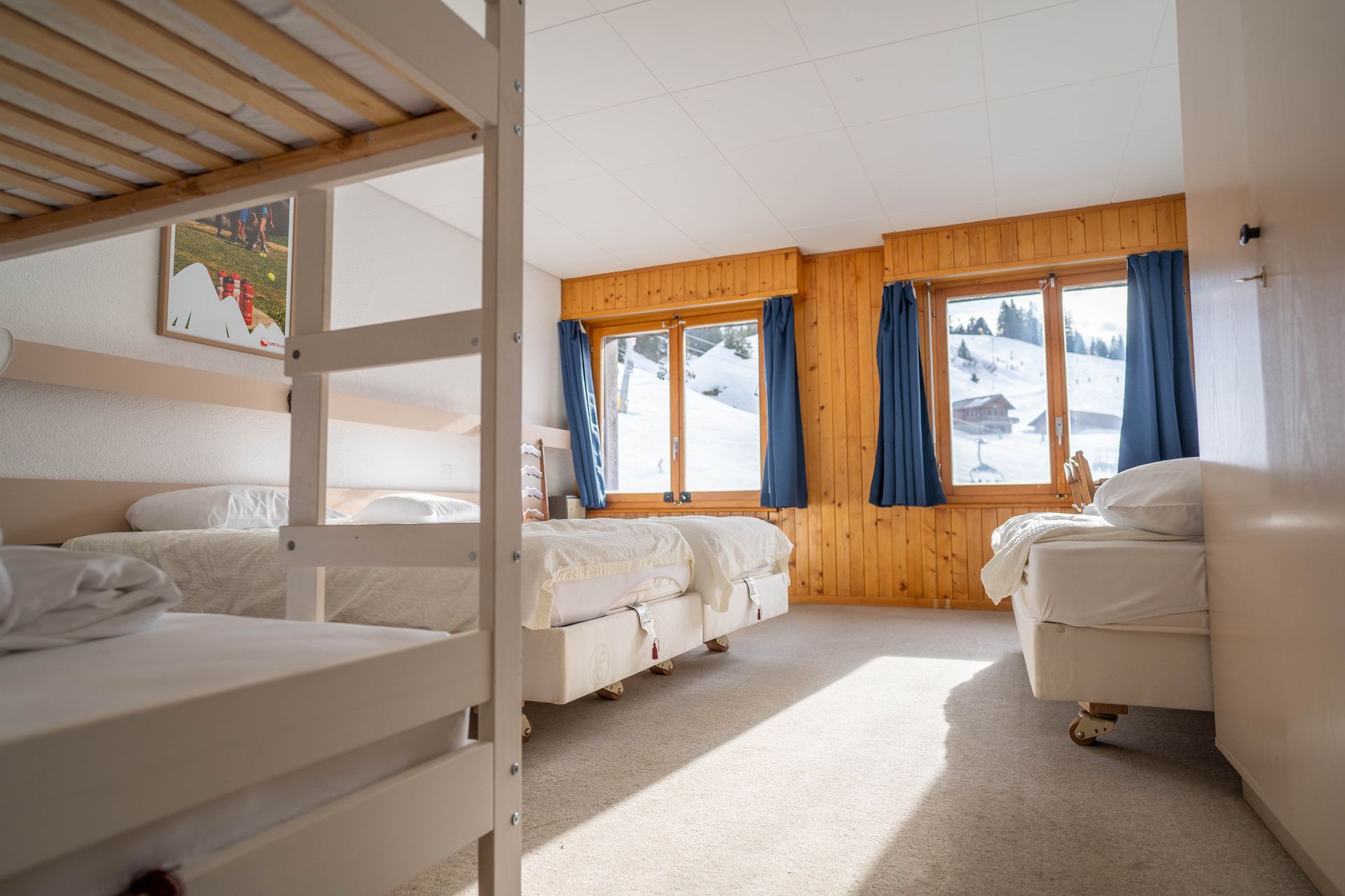 A brightly lit room with bunk beds, and three single beds with a snowy mountain view outside the windows.