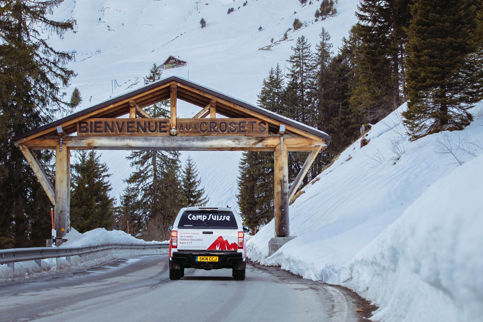 A pickup truck drives under a wooden archway in the snowy French Alps,