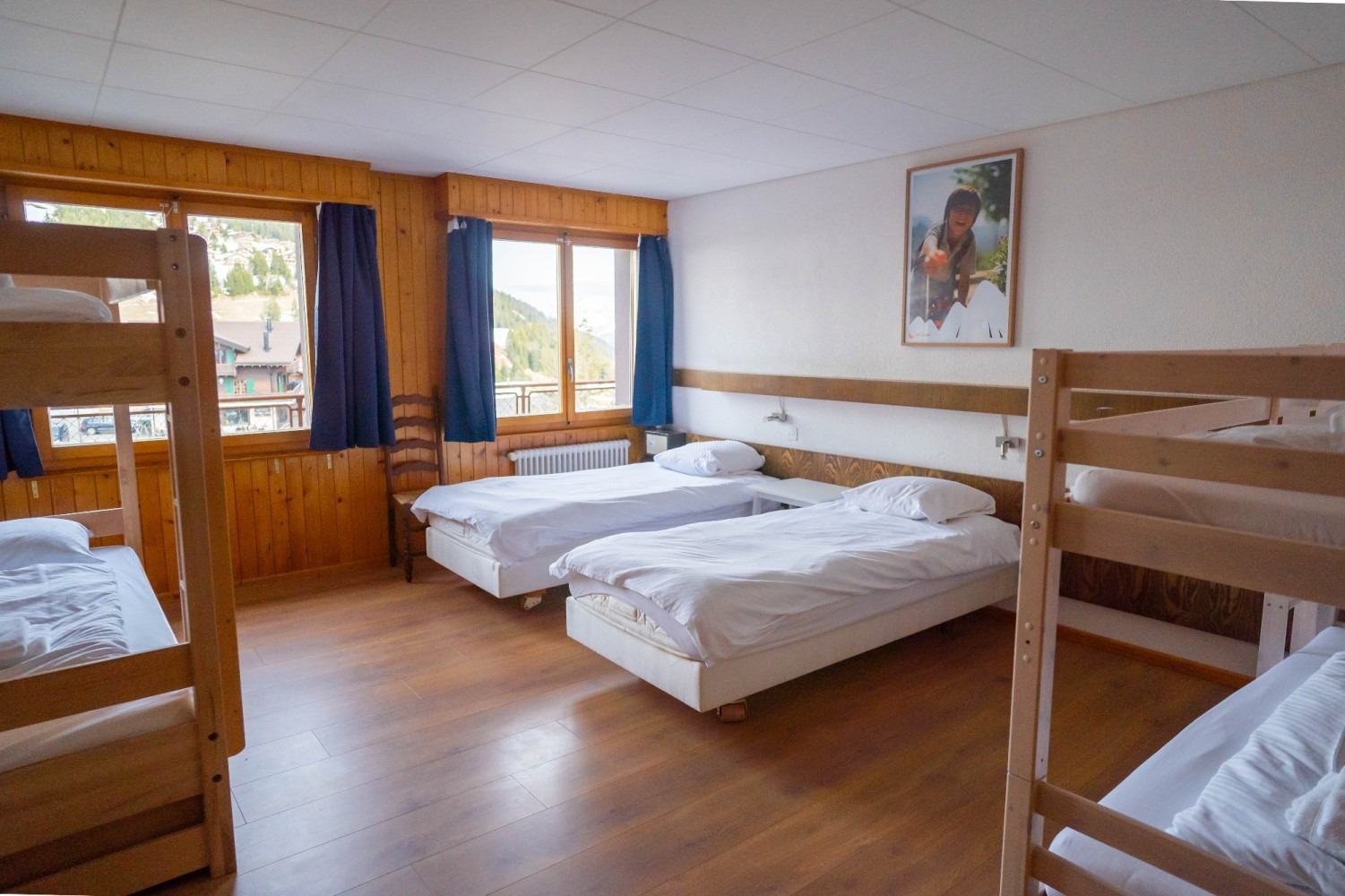 A dorm room with three bunk beds and two single beds, wooden walls, and natural light from windows.