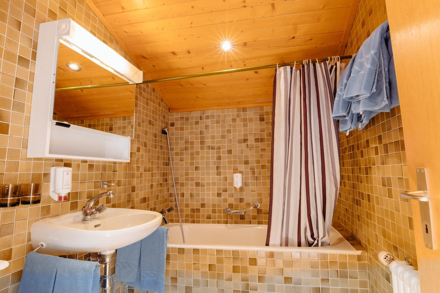 Bathroom with beige tiled walls, wooden ceiling, sink, bathtub, shower curtain, and towels.