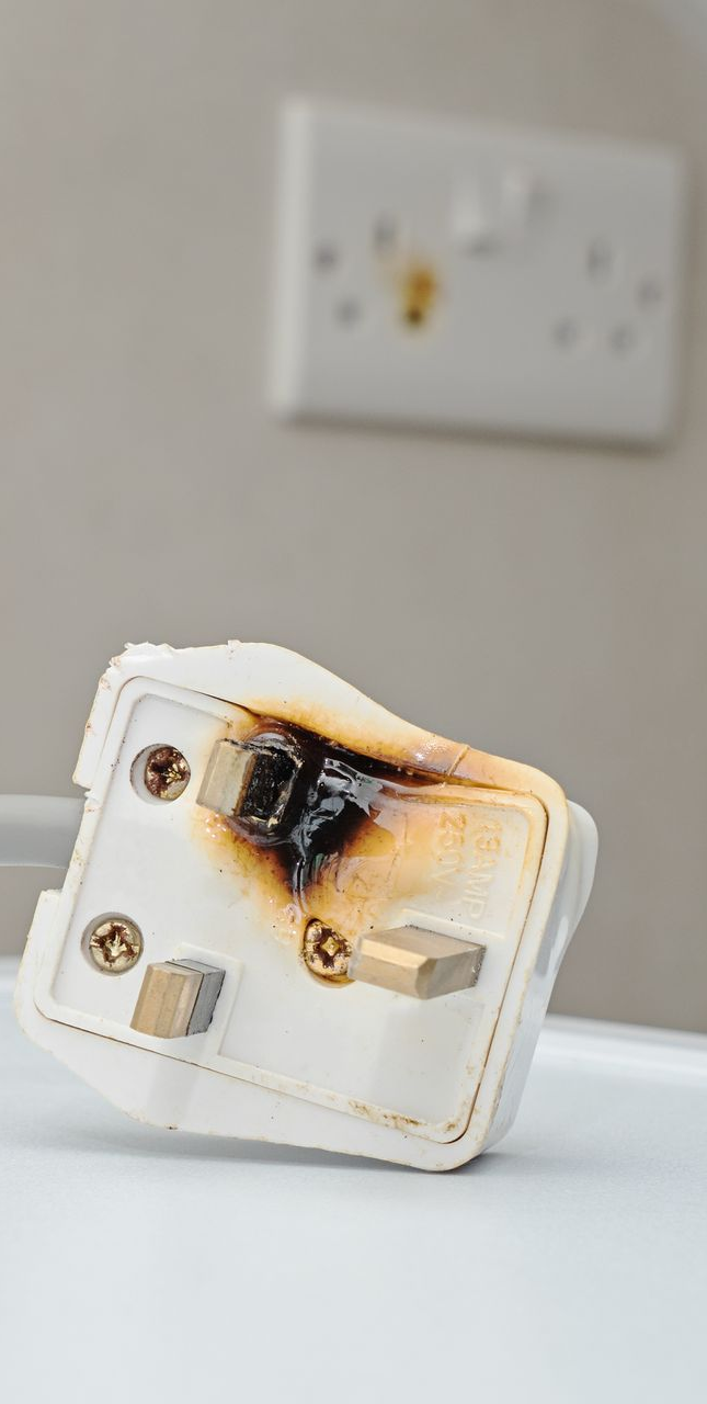 Burnt electrical plug lying in front of a wall outlet; charred plastic and metal.