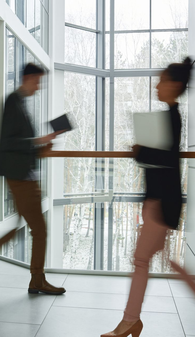 People walking and holding items inside a building with large windows.