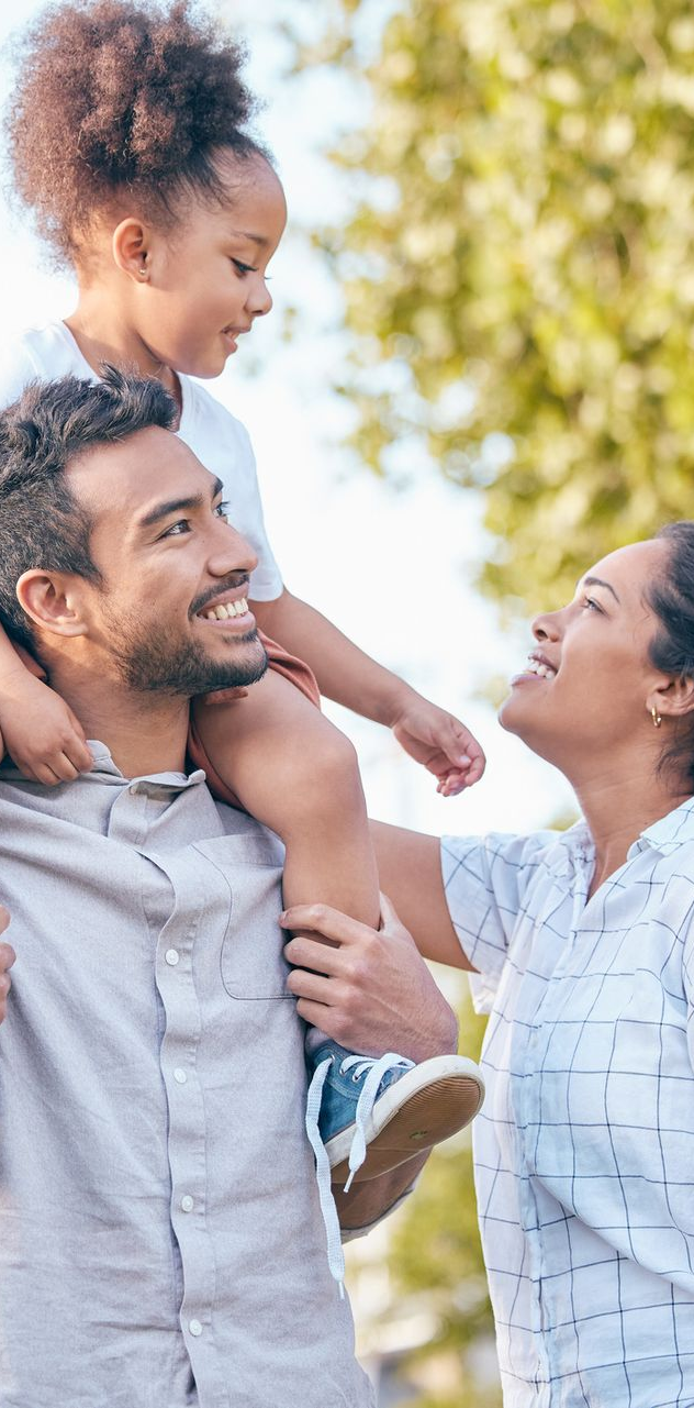 Family smiles while the father carries the child on his shoulders. The mother looks at the child. Outdoors.