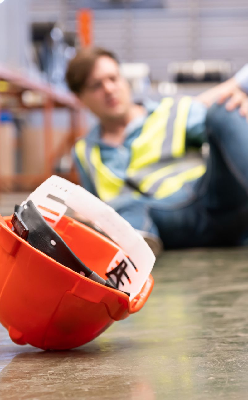 Worker lies on floor with orange hard hat and tools nearby. Yellow safety vest visible.
